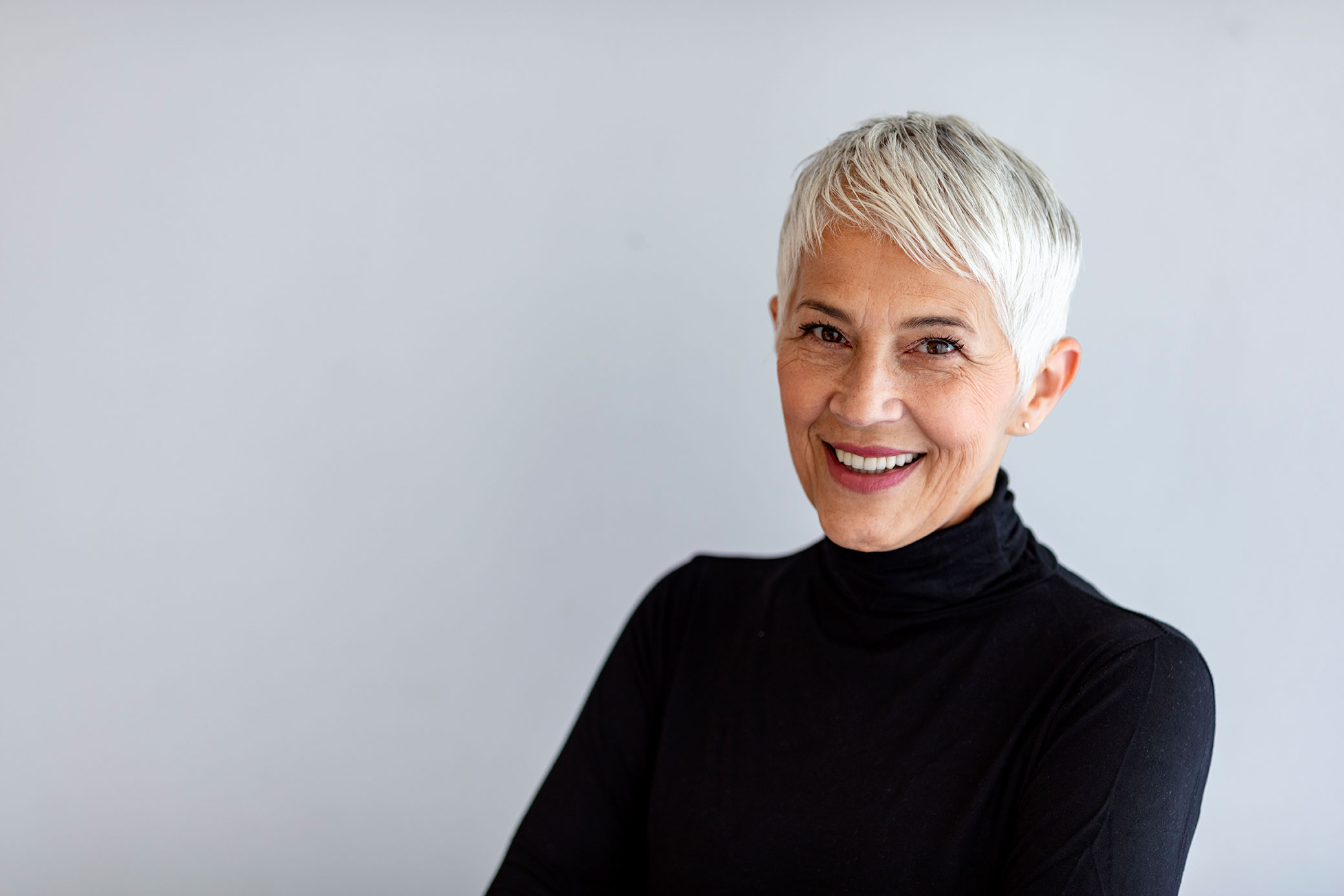 Close up portrait of beautiful older gray hair woman smiling and standing by gray wall wearing black turtleneck.
