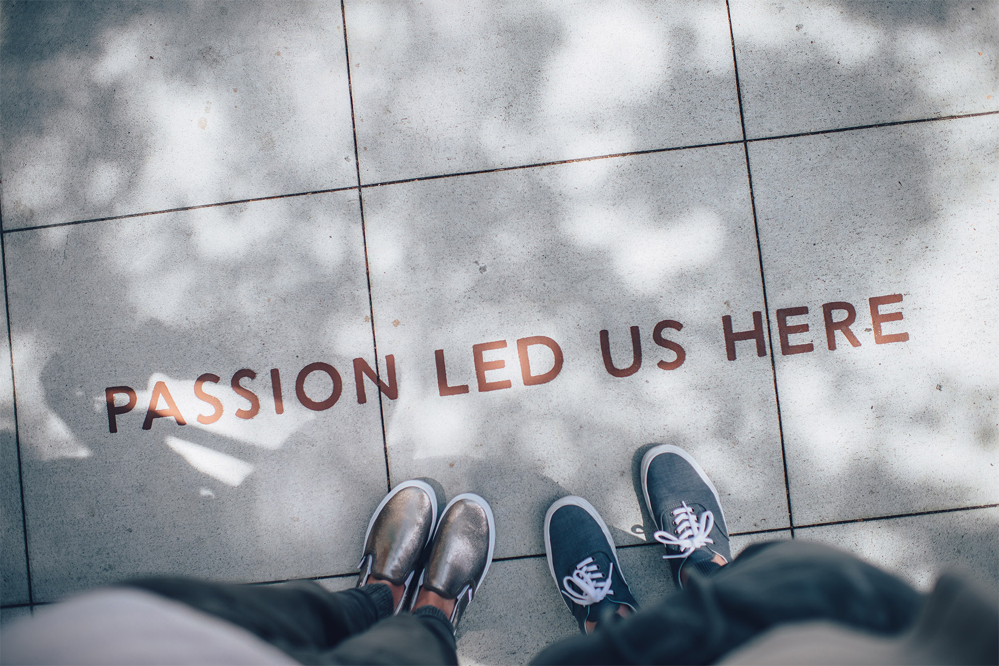 Aerial view of a sidewalk with the words passion led us here painted on the sidewalk. Two people's shoes and legs are in the shot as if they're the ones that took the photo.