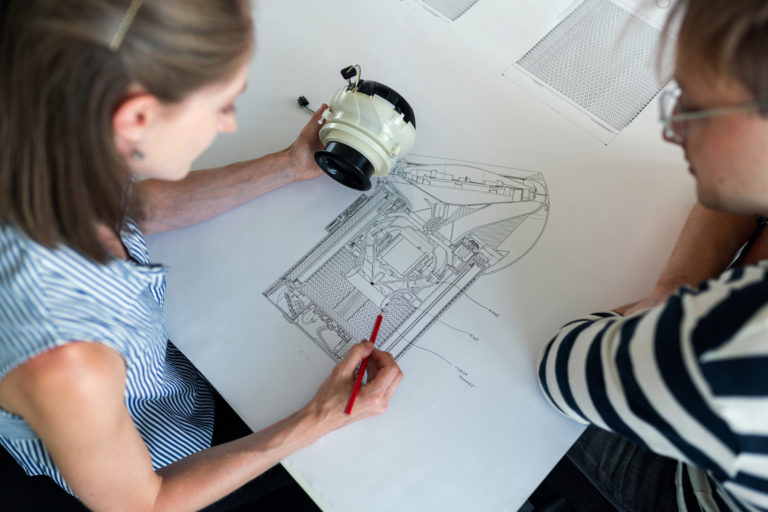 One employee drawing a diagram of a piece of equipment that she is holding in her hand while another is sitting on the side looking at the drawing.